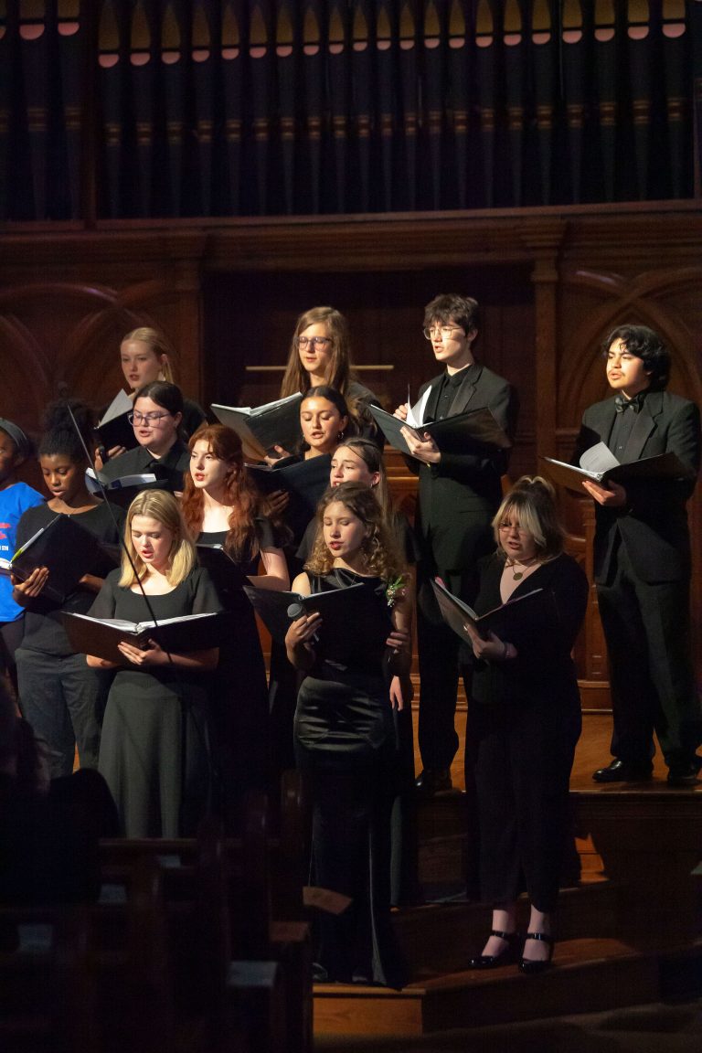 Teen singers in black concert dress hold folders and sing on the steps of a church sanctuary beneath organ pipes