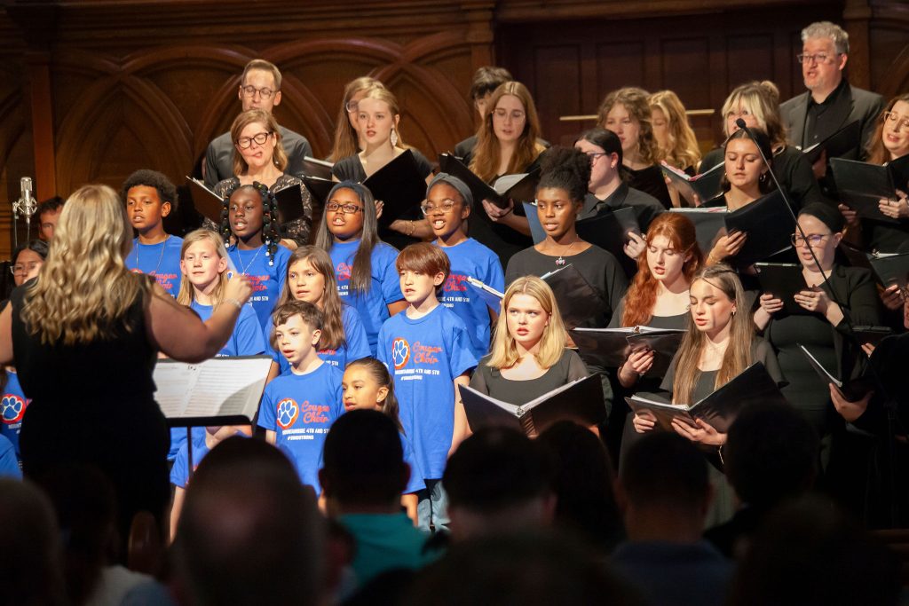 A large combined choir of children in bright blue shirts and adults in black sings in a wood-paneled church as a conductor leads