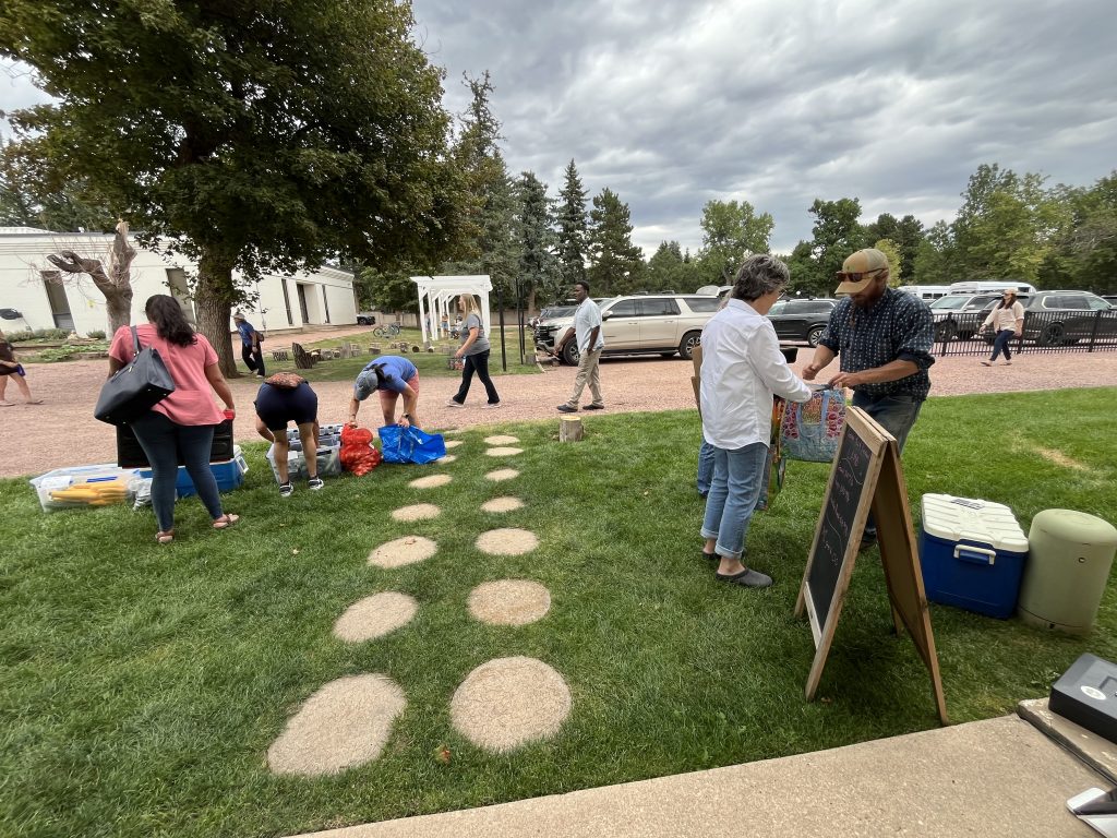 People sort bags and boxes of produce on a lawn beneath a large tree outside a white school building