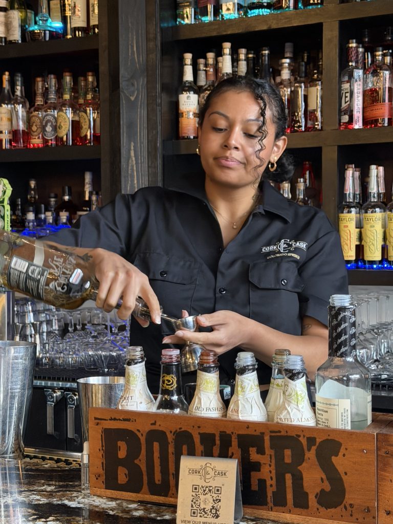 A bartender in a black Cork & Cask shirt pours a spirit into a jigger behind a bar lined with bitters bottles