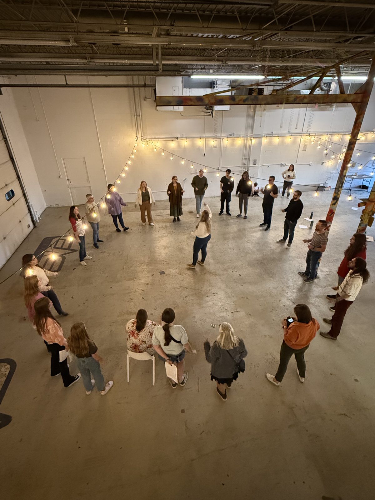 Seen from above, about two dozen singers stand in a wide circle on the concrete floor of a warehouse space strung with lights