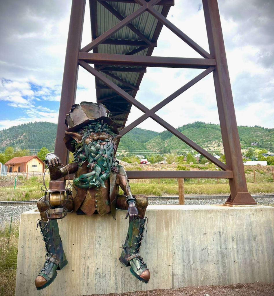 A steel troll sculpture with a green patina beard, a floppy hat and laced boots sits on a concrete footing beneath Palmer Lake's rust-colored pedestrian bridge, holding a lantern