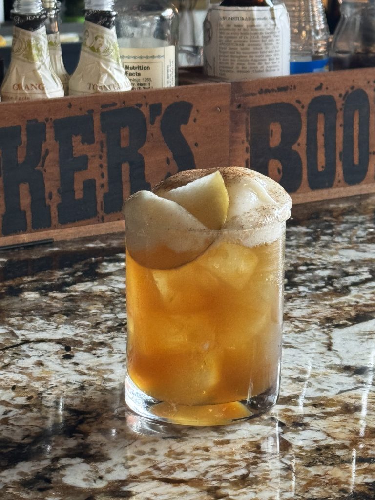 An amber Pear Potions cocktail in a rocks glass, topped with cinnamon-dusted foam and a slice of pear, on a granite bar